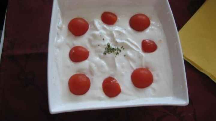 Eine wei&szlig;e Sch&uuml;ssel mit Zwiebelschmand, garniert mit Kirschtomaten und Kr&auml;utern, steht auf einem dunklen Tischtuch.