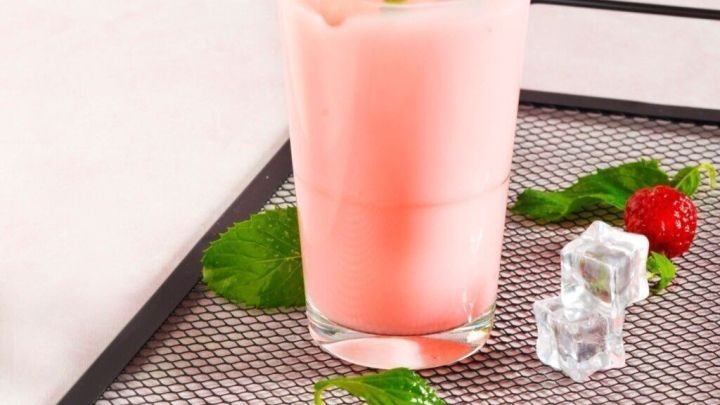 Ein gro&szlig;es Glas Himbeerlassi mit Pfefferminz liegt auf einem schwarzen Gittertablett, umgeben von frischen Himbeeren und Eisw&uuml;rfeln.