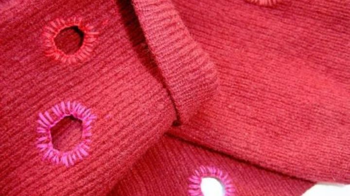 Roter Pulli mit farbigen gestickten L&ouml;chern, als Zeichen des Recyclings, liegt auf einem Tisch in heller Umgebung.
