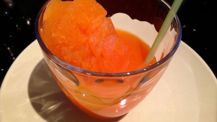 Ein Glas mit orangenem Sorbet steht auf einem Teller, umgeben von einem dunklen Tisch. Ein Strohhalm steckt hinein.