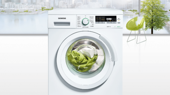 Waschmaschine w&auml;scht gr&uuml;ne Kleidung in modernem, hellem Raum mit Blick auf eine Stadtlandschaft und einem gr&uuml;nen Stuhl.