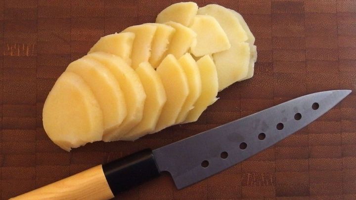 Gepellte Kartoffeln liegen auf einem Holzbrett, w&auml;hrend ein scharfer Messer geschnittene Scheiben daneben pr&auml;sentiert.