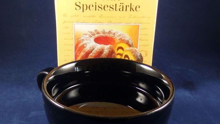 Eine schwarze Sch&uuml;ssel mit feiner Speisest&auml;rke steht vor einer Packung, die das Produkt zeigt, auf einem blauen Untergrund.