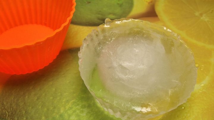 Ein transparentes, unzerbrechbares Eisw&uuml;rfelf&ouml;rmchen mit einem gro&szlig;en Eisw&uuml;rfel steht auf einer bunten Unterlage, neben einem orangefarbenen F&ouml;rmchen.