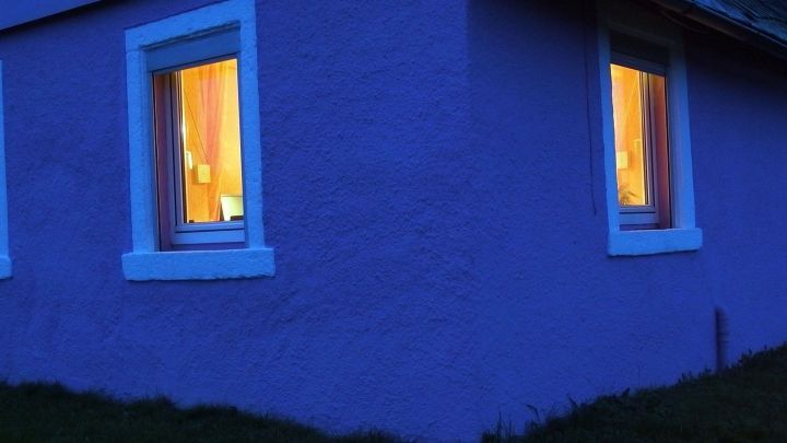 Beleuchtete Fenster eines Hauses strahlen warmes Licht aus, w&auml;hrend es in der D&auml;mmerung ruhig neben einer Wiese steht.