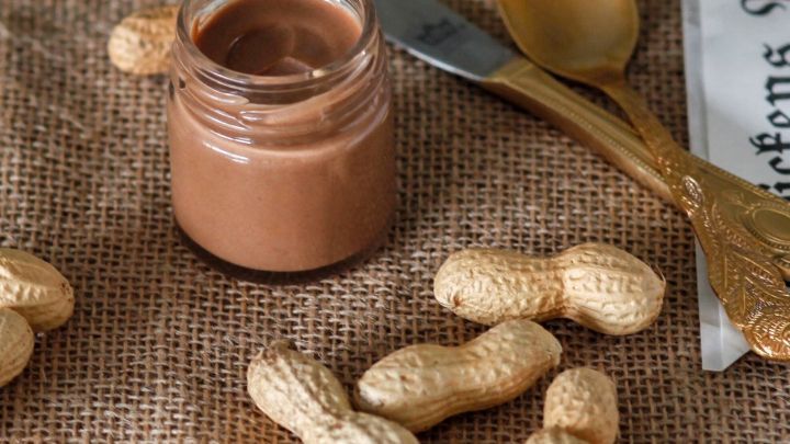 Ein Glas mit selbstgemachter Erdnussbutter steht auf einem juten Tuch, umgeben von ungesch&auml;lten Erdn&uuml;ssen und Besteck.