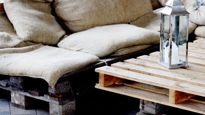 Ein gem&uuml;tliches Sofa aus Europaletten mit beigen Kissen steht neben einem Holztisch und einer Laterne in einem Innenraum.