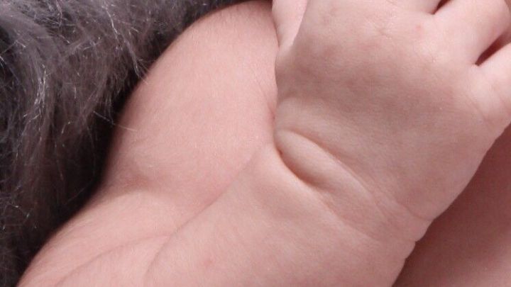 Ein Babyarm mit zarter Haut greift spielerisch in den Mund, umgeben von einer weichen, grauen Unterlage.