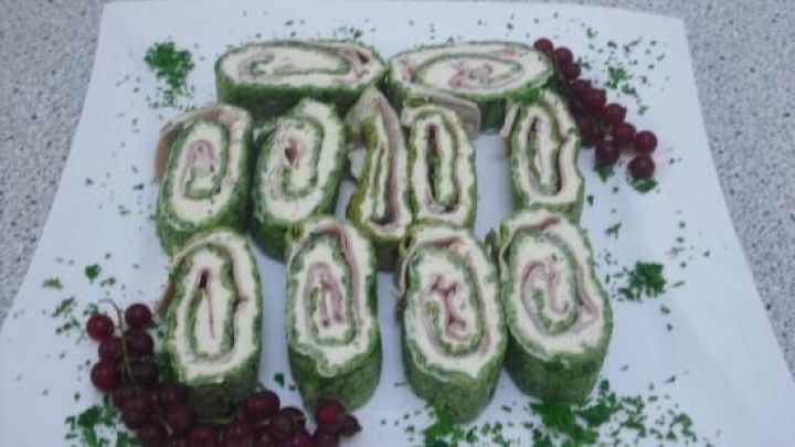 Spinatrollen mit Frischk&auml;se und Schinken, dekorativ auf einem wei&szlig;en Teller platziert, umgeben von frischen Kr&auml;utern und roten Beeren.