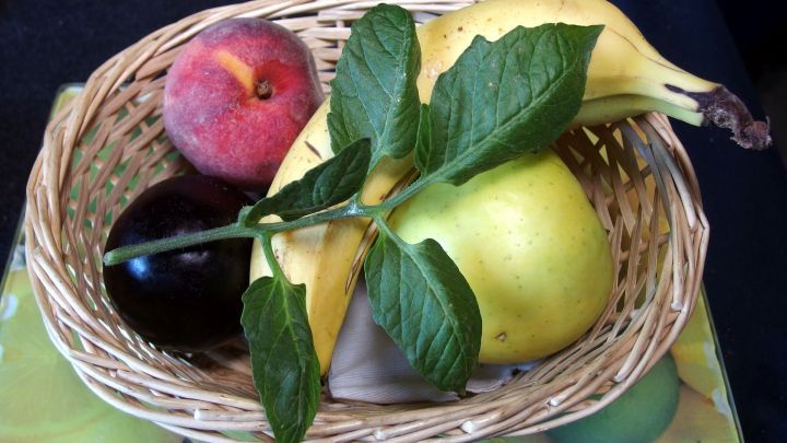 In einem geflochtenen Korb liegen frische Fr&uuml;chte wie &Auml;pfel, Bananen und eine Birne, umgeben von gr&uuml;nen Bl&auml;ttern.