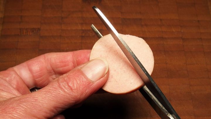 Eine Hand h&auml;lt eine K&uuml;chenschere, die eine runde Scheibe durchschneidet, auf einem holzfarbenen Tisch.