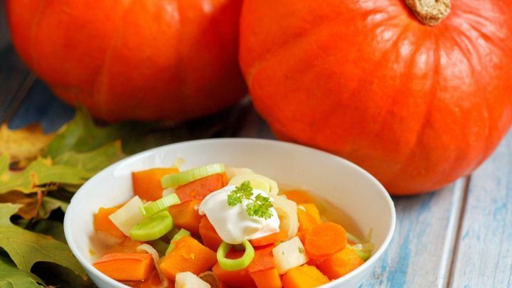 Hokkaido-K&uuml;rbissuppe in wei&szlig;er Schale, garniert mit Sauerrahm und Kr&auml;utern, umgeben von frischen K&uuml;rbissen auf Holztisch.