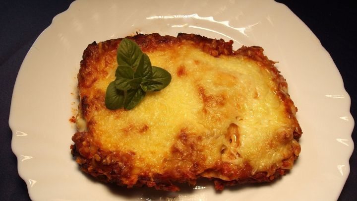 Ein St&uuml;ck Lasagne mit geschmolzenem K&auml;se liegt auf einem wei&szlig;en Teller, dekoriert mit einem frischen Basilikumblatt.