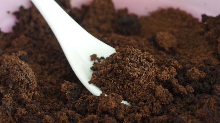 Ein wei&szlig;er L&ouml;ffel greift in ein Sch&auml;lchen mit dunklem Kaffeepeeling, das auf einem rosa Untergrund liegt.