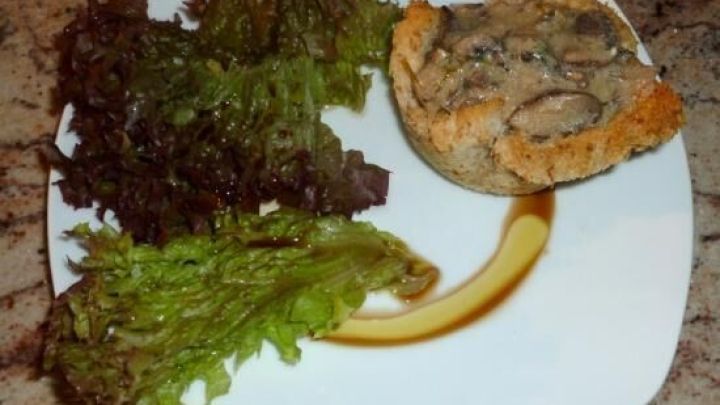 Ein K&ouml;rbchen aus Brot liegt gef&uuml;llt mit Pilzcreme auf einem Teller, flankiert von frischen gr&uuml;nen Salatbl&auml;ttern.