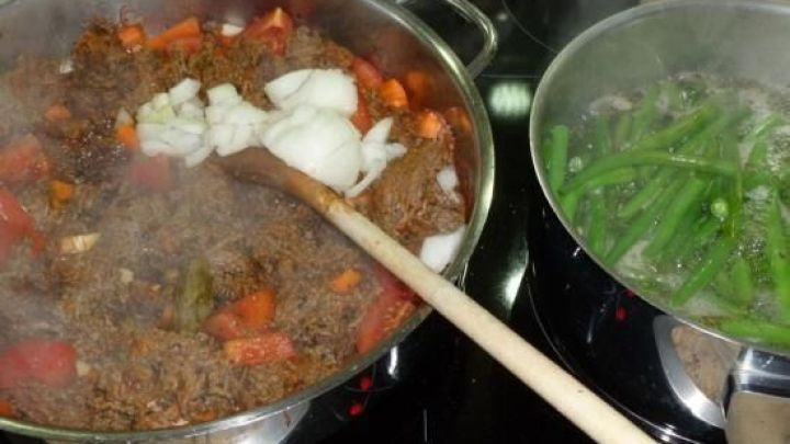 Scharf gew&uuml;rztes Rindfleischhack brutzelt in einer Pfanne, w&auml;hrend daneben geschmorte Stangenbohnen dampfen.