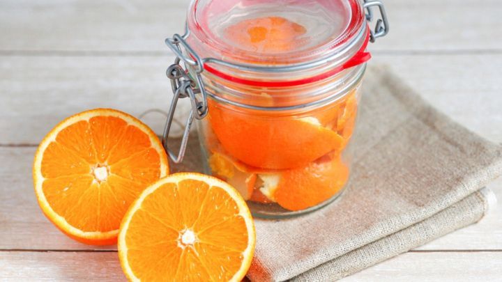 Ein Glas mit kandierten Orangenschalen steht auf einem Stoff unterhalb zweier halbierter Orangen in einer hellen, einladenden Umgebung.