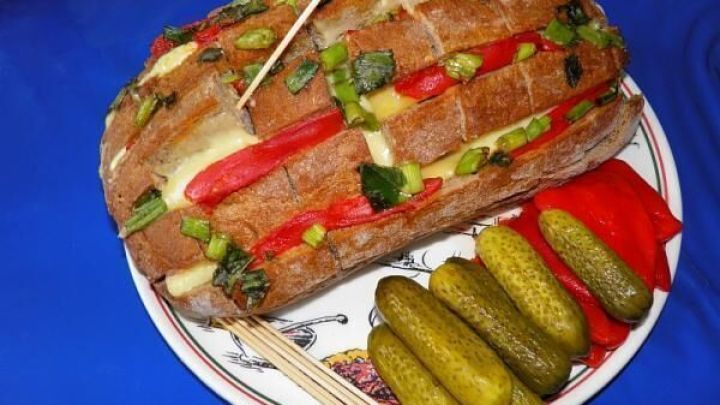 Ein Brotlaib, mit geschmolzenem K&auml;se, Paprika und Lauch gef&uuml;llt, liegt auf einem Teller mit Gurken und Paprikascheiben.