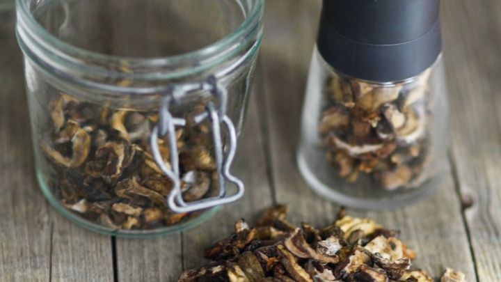 Getrocknete Steinpilze liegen verstreut auf einem Holztisch, neben einem offenen Glas und einem Gew&uuml;rzstreuer.