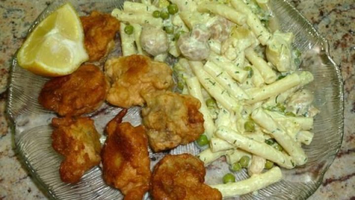 Auf einem Teller liegen goldene, knusprig ausgebackene Champignons im Hefe-Bierteig, umgeben von Nudel-Salat und einer Zitronenscheibe.