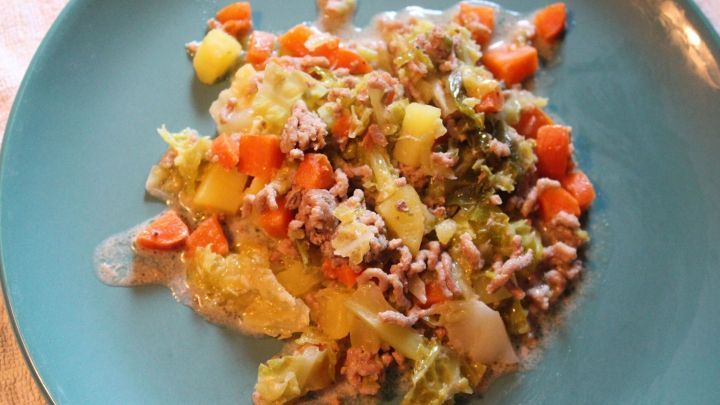 Ein bunter Wirsingeintopf mit Hackfleisch liegt auf einem blauen Teller, umgeben von einem rustikalen Tisch.