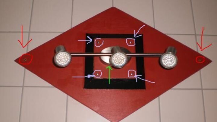 Eine Badlampe mit drei Strahlern ist mittig an einer rot-schwarzen rautenf&ouml;rmigen Platte auf Fliesen befestigt.