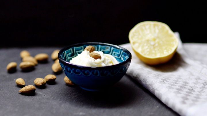 Eine kleine Sch&uuml;ssel mit Mandel-Mayonnaise steht auf einem dunklen Untergrund, umgeben von Mandeln und einer halbierten Zitrone.