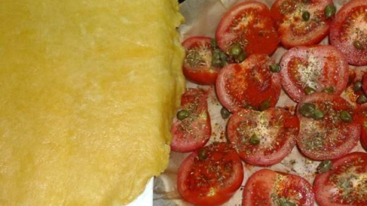 Eine knusprige Teigbasis liegt neben frischen, gew&uuml;rzten Tomatenscheiben auf Backpapier, bereit f&uuml;r die Zubereitung.