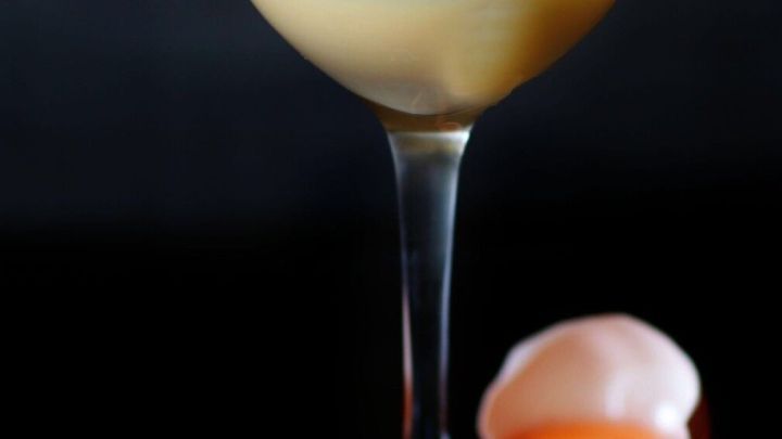 Ein Glaskelch mit Eierlik&ouml;r steht auf einer dunklen Fl&auml;che, neben einem Eissw&uuml;rfel und einem frischen Eigelb.