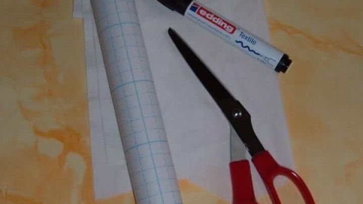 Ein gerolltes Blatt Papier steht neben einer Schere, einem Marker und einem weiteren St&uuml;ck Stoff auf einem hellen Untergrund.