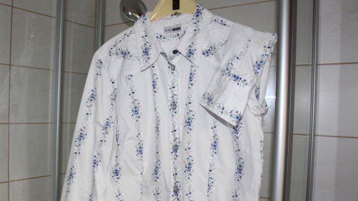 Ein wei&szlig;es, gebl&uuml;mtes Hemd h&auml;ngt an einem B&uuml;gel im Bad, umgeben von gefliesten W&auml;nden und einer Duschkabine.