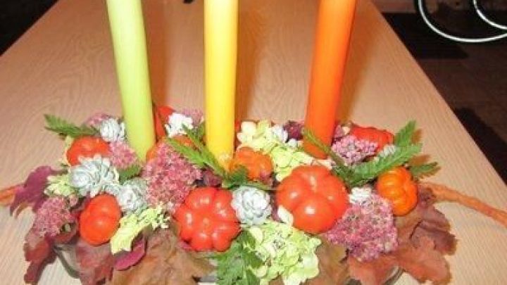 Ein dekorativer Herbsttisch mit drei bunten Kerzen in Gelb und Orange, umgeben von K&uuml;rbissen und buntem Laub.