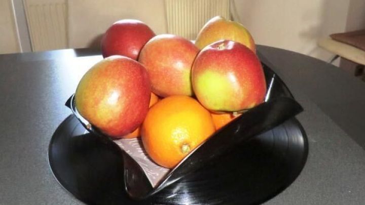 Eine Obstschale aus Schallplatte h&auml;lt mehrere &Auml;pfel und eine Orange auf einem Tisch in einem hellen Raum.