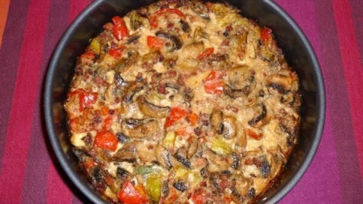 Eine frisch gebackene Champignon-Schinken-Torte aus einer runden Form, umgeben von buntem Gem&uuml;sestreifen auf einem Tisch.