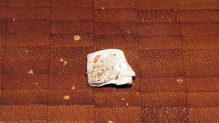 Ein Kaugummist&uuml;ck liegt auf einem Holzuntergrund, umgeben von Brotkr&uuml;meln, die darauf hinweisen, dass gegessen wurde.