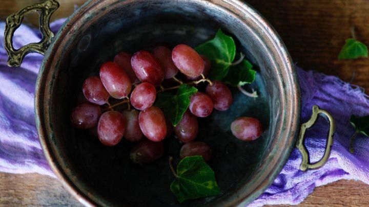 rote Weintrauben liegen in einer Sch&uuml;ssel mit Wasser, um gereinigt zu werden, auf einem Holzuntergrund mit lila Tuch.