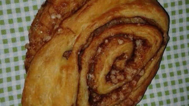 Ein frisch gebackenes Franzbr&ouml;tchen liegt auf einem karierten Tuch, zeigt seine spiralf&ouml;rmige F&uuml;llung aus Zimt und Zucker.