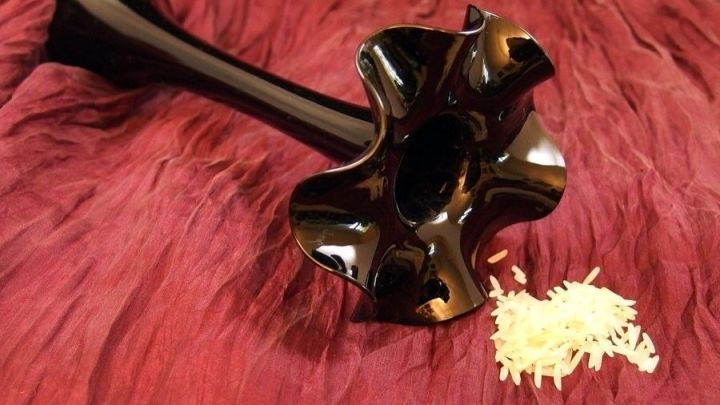 Eine schmale, schwarze Vase liegt auf einem weichen, roten Untergrund, daneben liegen verstreute Reisk&ouml;rner.