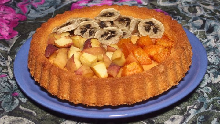 Ein runder Kuchenteig f&uuml;r Obst, belegt mit frischen Apfel- und Aprikosenscheiben, steht auf einem blauen Teller.