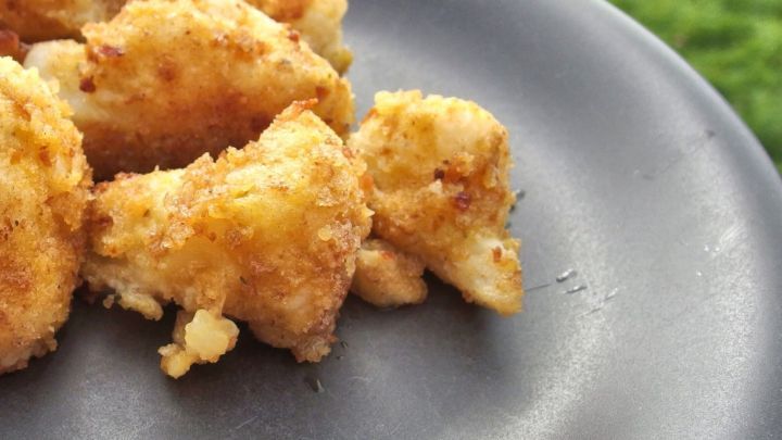 Panierte Blumenkohlr&ouml;schen liegen knusprig auf einem dunklen Teller, umgeben von gr&uuml;nem Gras im Hintergrund.