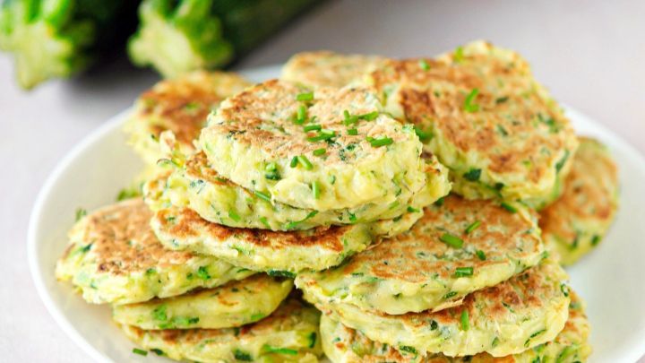 Zucchini-Pfannkuchen liegen auf einem Teller und werden von frischen Zucchini im Hintergrund umgeben.