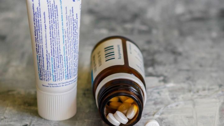 Ein R&ouml;hrchen mit einer Creme und ein offenes Fl&auml;schchen mit wei&szlig;en Tabletten liegen auf einer grauen Oberfl&auml;che.