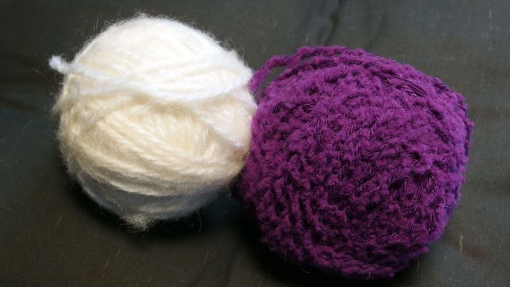 Zwei Wollreste, eine wei&szlig;e und eine lila, liegen auf einem dunklen Untergrund und warten darauf, verarbeitet zu werden.