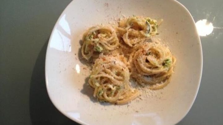 Pasta liegt gerollt auf einem wei&szlig;en Teller mit geriebenem K&auml;se und Zucchini. Die Umgebung scheint eine moderne K&uuml;che zu sein.
