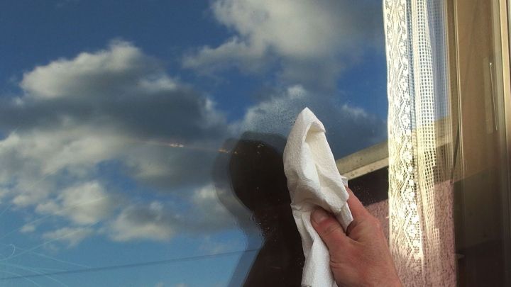 Eine Hand putzt eine Fensterscheibe mit einem Tuch, w&auml;hrend die Wolken am blauen Himmel reflektiert werden.