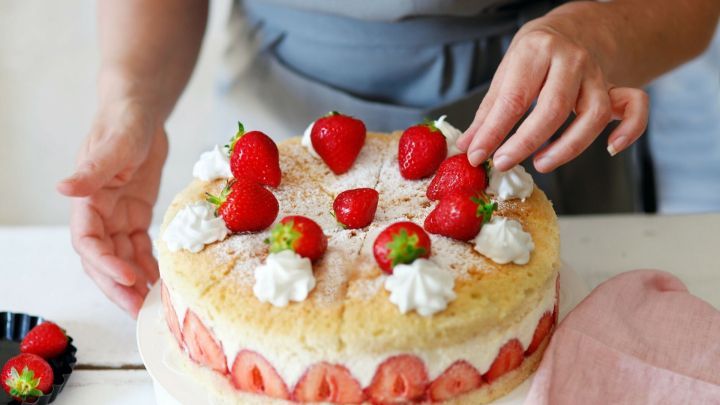 Ein Tortenboden wird sorgf&auml;ltig mit frischen Erdbeeren und Sahne dekoriert, auf einem hellen Tisch in einer K&uuml;che.