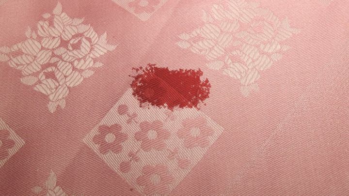 Ein roter Blutfleck befindet sich auf einem zart rosa, gemusterten Stoff. Der Fleck ist deutlich sichtbar.