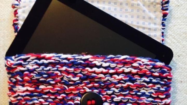 Eine bunte, gestrickte Tablet-Tasche liegt offen da, w&auml;hrend ein schwarzes Tablet darin fast herausragt.