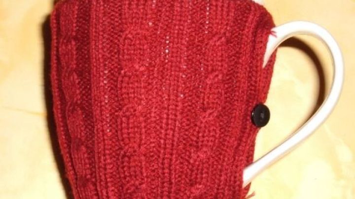 Eine rote Strickh&uuml;lle umschlie&szlig;t eine wei&szlig;e Kaffeetasse, die auf einem hellen Untergrund steht und W&auml;rme spendet.