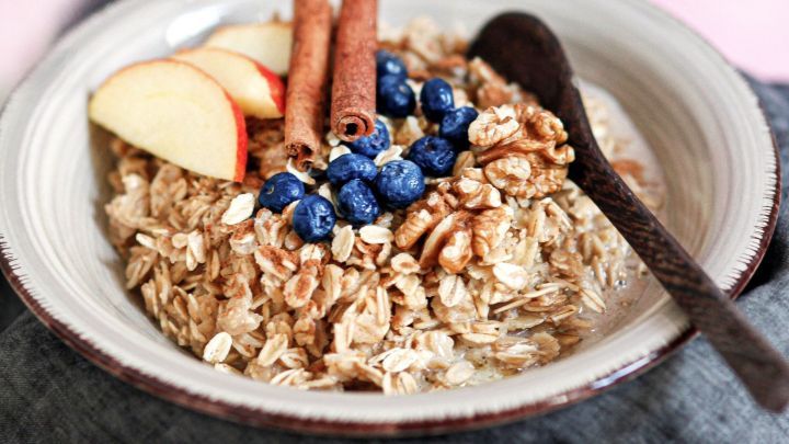 Haferbrei wird in einer Sch&uuml;ssel mit Zimtstangen, &Auml;pfeln und Blaubeeren dekoriert, umgeben von einem grauen Tuch.